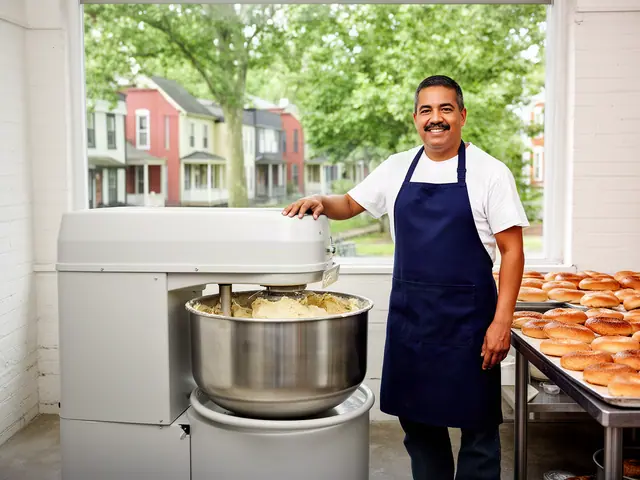 A smiling baker in a navy apron beside a new gray spiral mixer with a bowl of dough and fresh bagels.
